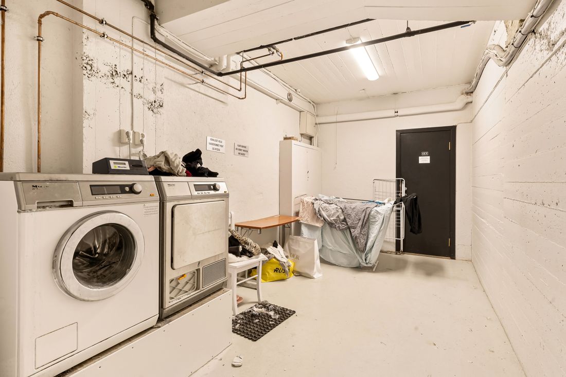 A windowless basement laundry with a washing machine and a tumble dryer side by side, exposed pipes overhead and wet washing spread over a drying rack