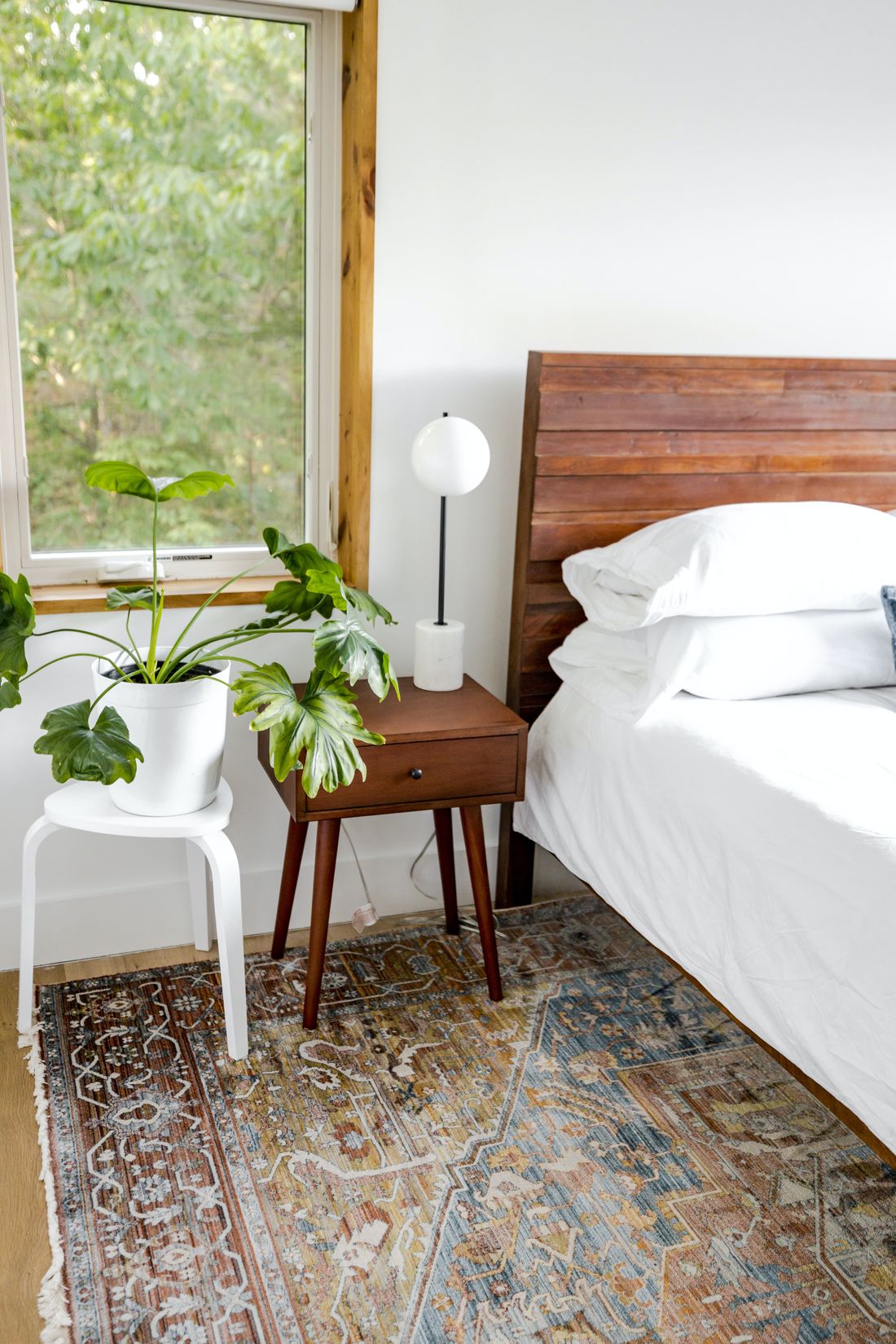 A bedroom corner with a philodendron on a stool right beside a large window, a nightstand and lamp next to it, and the bed to the right