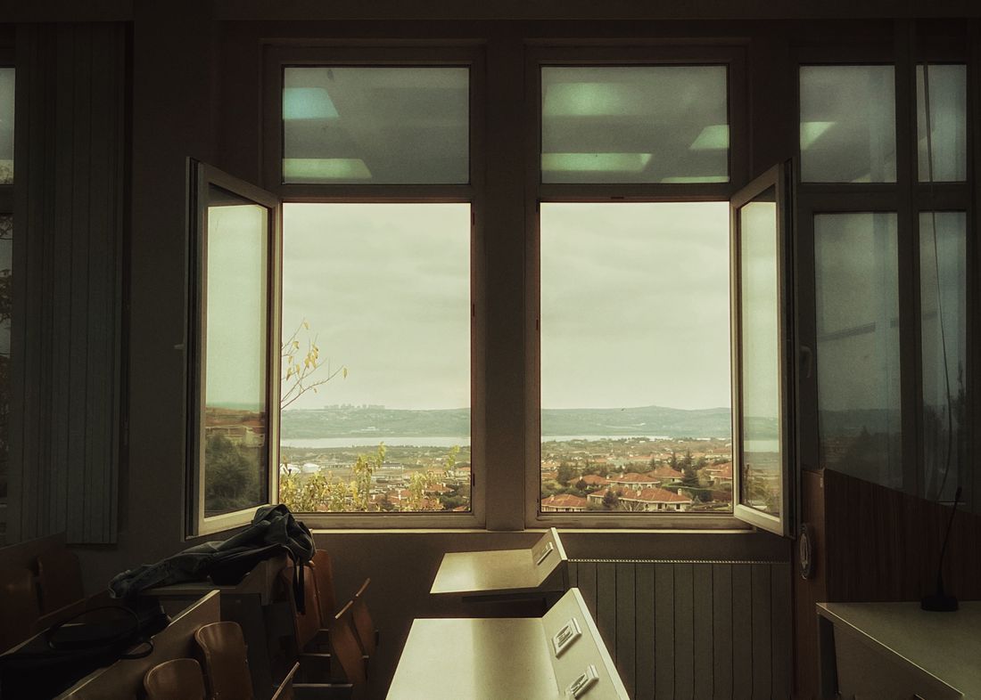 An empty classroom with two casement windows propped wide open above a radiator, chairs stacked and desks unoccupied