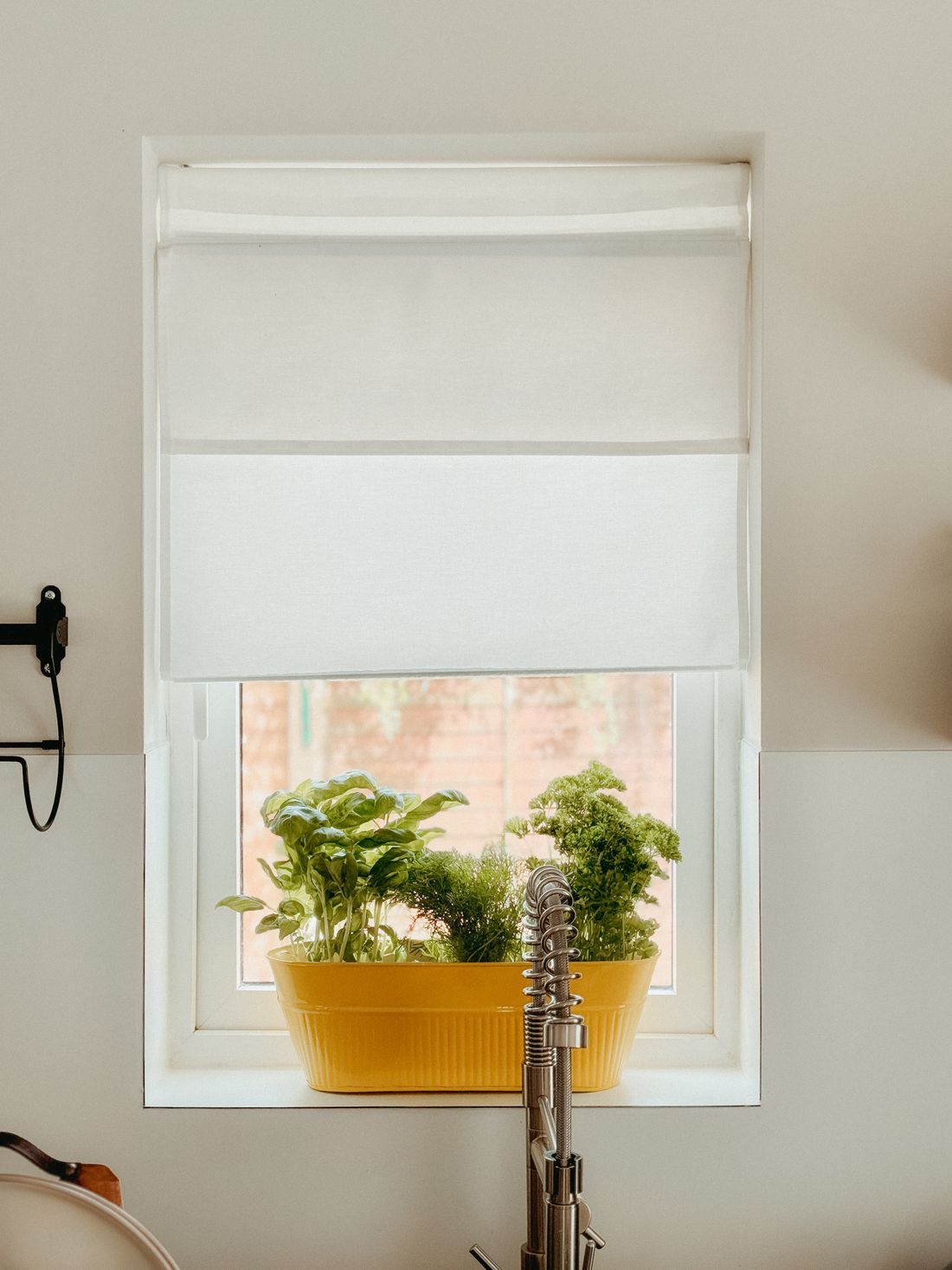 A yellow trough of basil, dill and parsley on a small kitchen windowsill above a sink, with a fabric blind pulled down over the top two thirds of the window