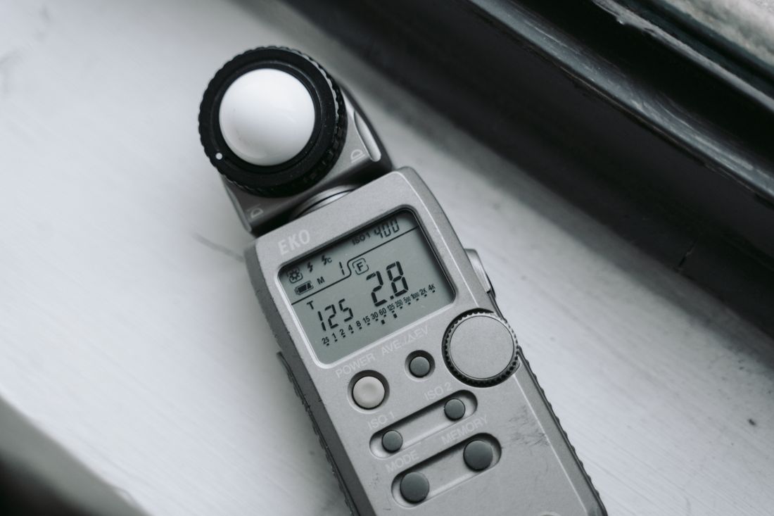 A handheld incident light meter on a windowsill, its display showing a shutter speed and an aperture