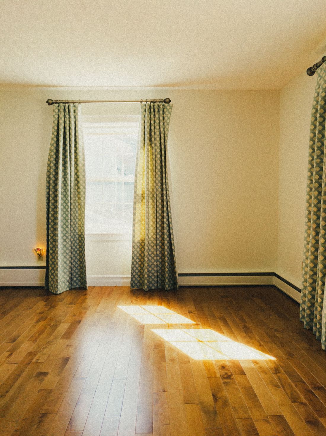 A curtained window in an empty room, one long bar of sunlight lying across a wooden floor and stopping about two metres in