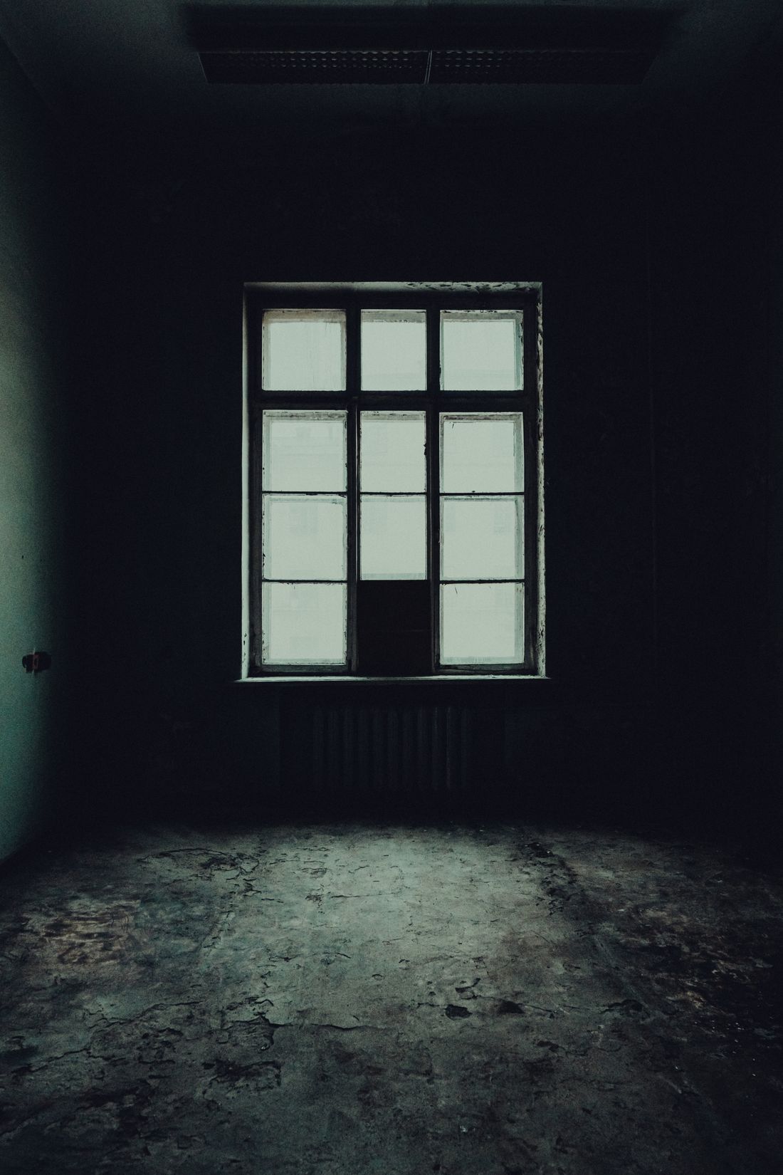 A bare, very dark room with a single multi-pane window above a radiator, the daylight reaching only a short way across the floor before the room goes black